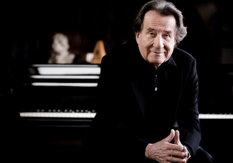 Rudolf Buchbinder sits with his hands touching, with a piano keyboard blurred out behind him.