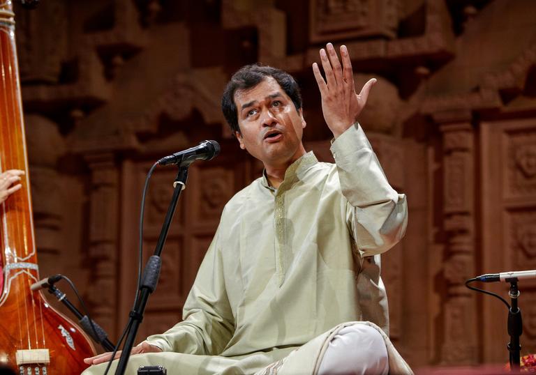 Uday Bhawalkar singing with his left hand raised. He is sitting cross-legged wearing a pale green tunic.