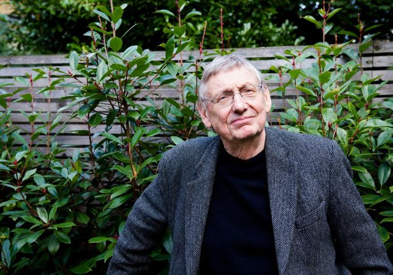 John Cumming wearing a dark suit, in front of a bush and fence