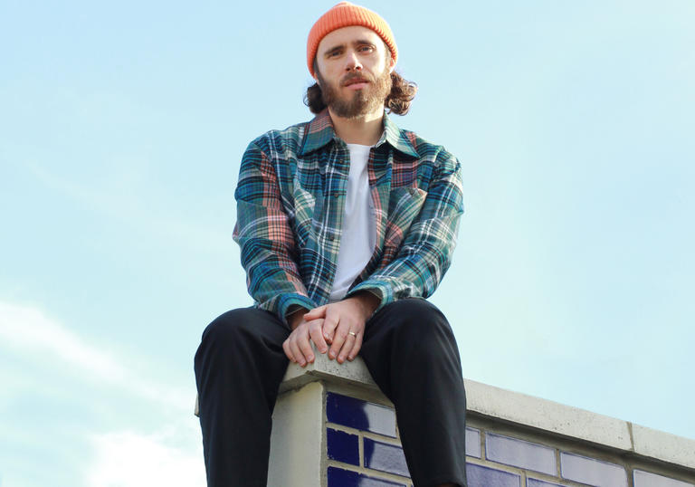 James Vincent McMorrow sitting on the corner of a tiled roof. He is wearing an orange beanie and checked shirt.