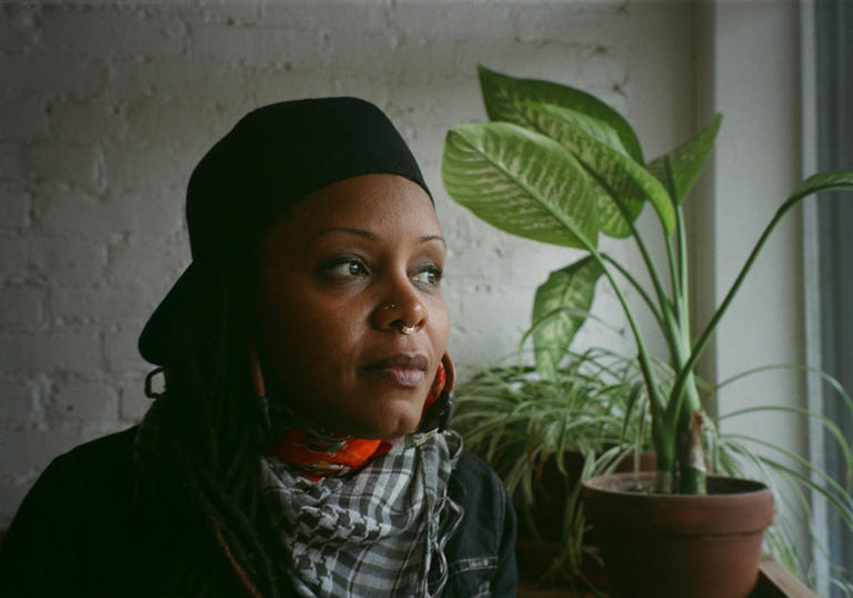 Matana Roberts looking out a window with a plant in front of her. She is wearing a black cap backwards and a white and black dogtooth scarf.