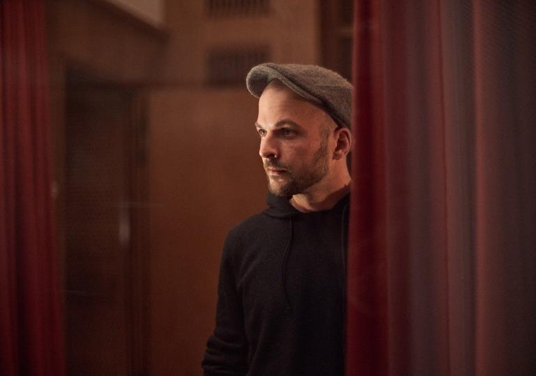 Nils Frahm standing between two red curtains, wearing a flat cap with a black hoodie