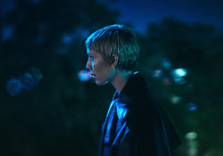 A night-time shot of Lykke Li standing side-on to the camera, wearing a leather jacket