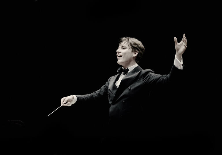 Klaus Makela is conducting in front of a black background, his right hand holding the baton and his left hand above his shoulder