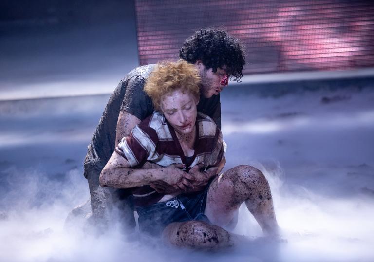 Two performers covered in mud and blood hold onto each other as smoke gathers around them.