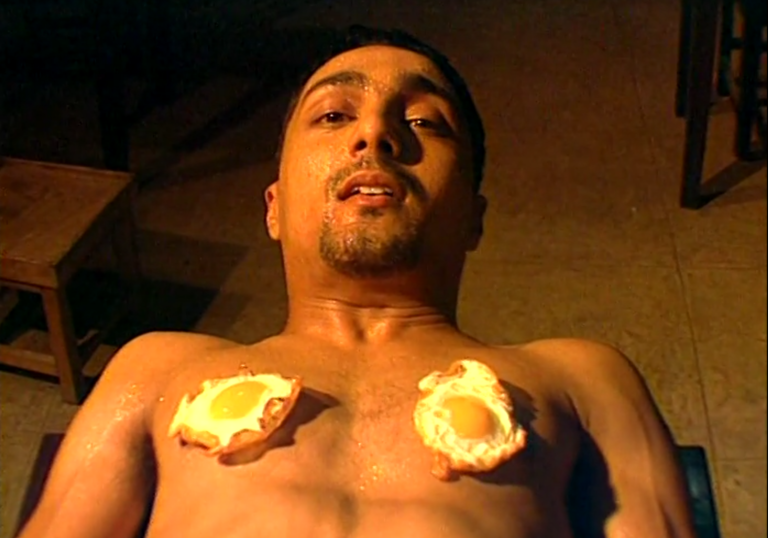 A still from Bomgay, a young shirtless man reclines with fried eggs on his chest