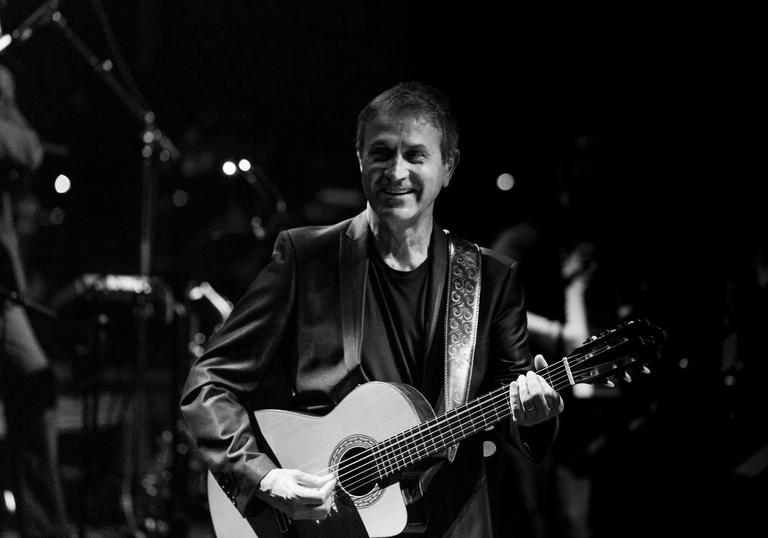 Black and white image of George Dalaras onstage holding a guitar smiling