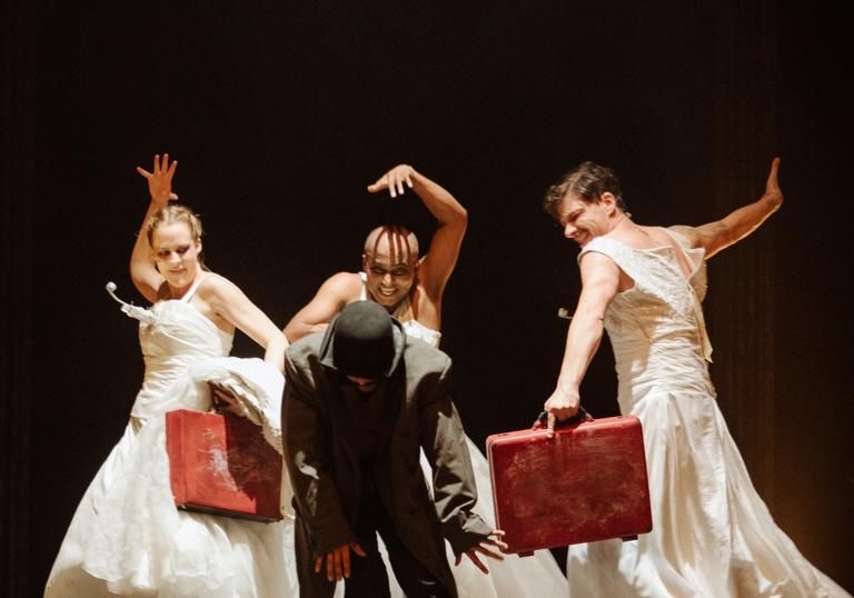 Three performers in white dresses appear to be dancing or moving behind a figure in a coat and hat in front of them. They each carry a red suitcase. 