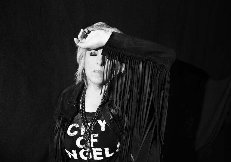 Black and white photo of Lucinda Williams wearing a black suede tassel jacket, arm aloft to her head