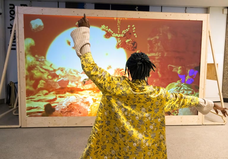 A person with their back to the camera standing in front of an interactive art installation, waving their hands above their head to prompt a visual response.