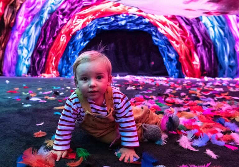 A baby crawls along a floor that is littered in different colourful fabrics in front of an arch made out of blue, purple and pink material.
