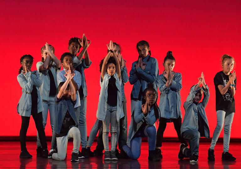 A group of children stand on stage in front of a red backdrop, dressed in denim, staggered in height with some holding their hands up over their heads.