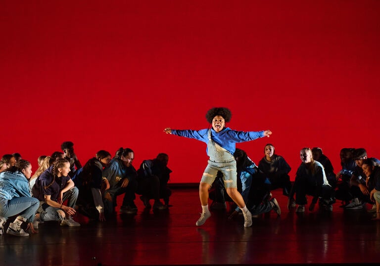 In front of a red background dancers are crouched on the floor and one dancer is in the air with their hands stretched out wearing a blue hoody
