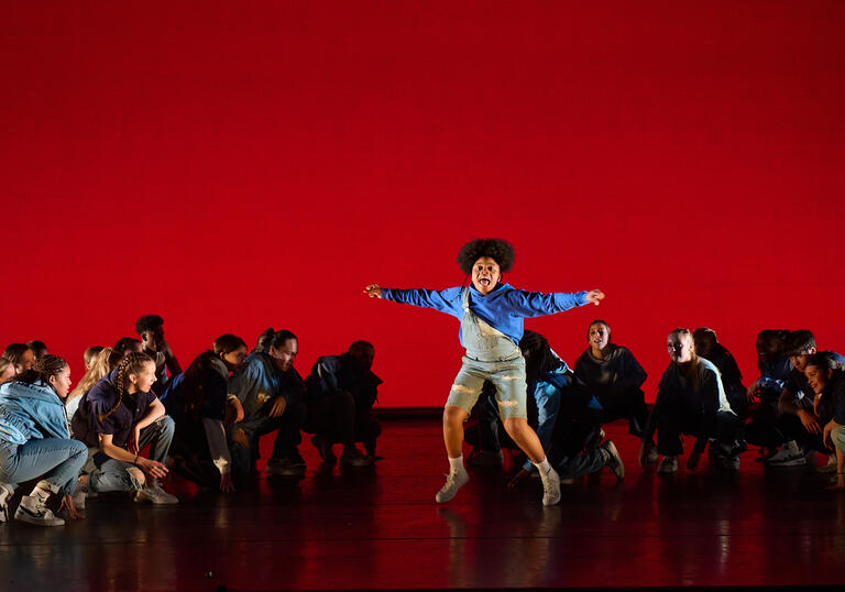 In front of a red background dancers are crouched on the floor and one dancer is in the air with their hands stretched out wearing a blue hoody