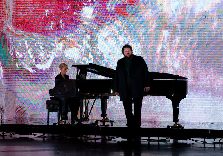 Allan Clayton stands in front of a piano at which Kate Golla sits, with a pink landscape by Fred Williams OBE projected on a screen behind them