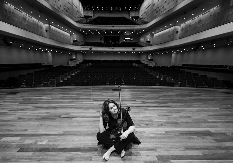 Patricia Kopatchinskaja sitting on a stage holding her violin in her lap