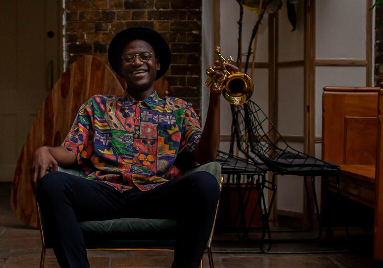 Mark Kavuma sitting laughing in a chair, laughing, holding a trumpet in a brightly patterened shirt and brown hat