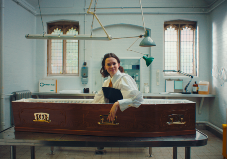 Person sits upright in coffin, camera from sideview.