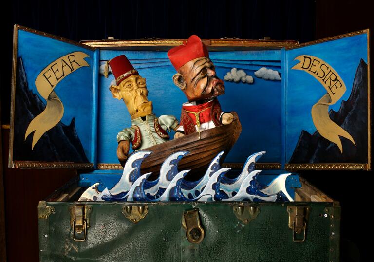 Two puppets in a travel case appear to be sailing on the sea in a boat. The words fear and desire are painted onto the back of the travel case on either side of them.
