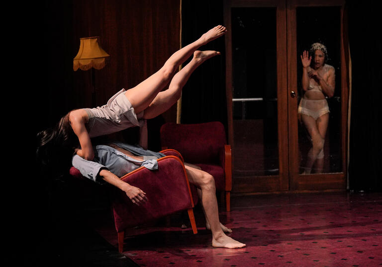A production photo of Peeping Tom performing. A woman stands at a glass door, looking in on a woman who levitates above a man sitting in a chair.