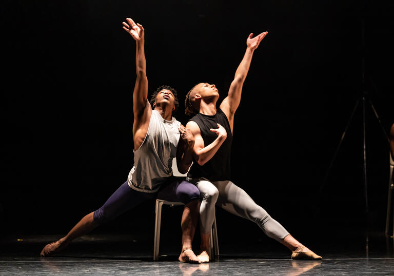 Two Ballet Black performers sit on the same chair with one hand raised towards the sky.