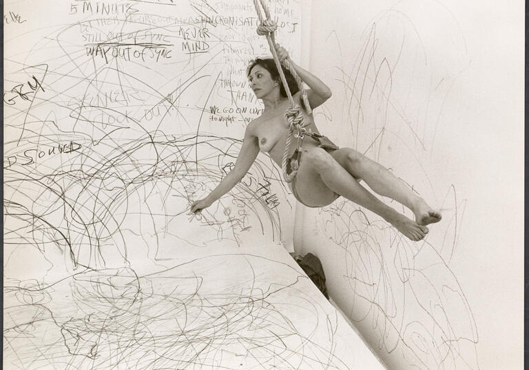 Image of a person without clothing swinging on a rope with a wall of drawings in the background