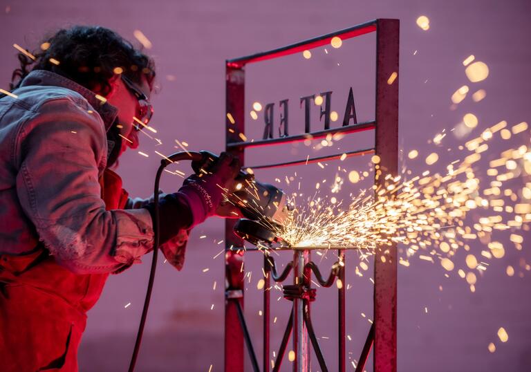 Rachel Mars welds a gate with the words 'After' shown backwards. Sparks are flying and they are set against a purple background