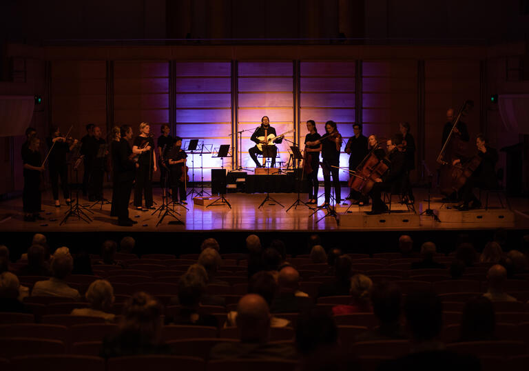 William Barton sits in the middle of the stage on a pedestal playing guitar and didgeridoo. Members of the ACO stand either side of him, in near-darkness