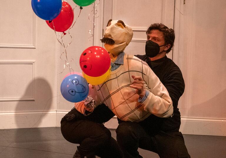 Bill the puppet kneels on the floor while holding a string of colourful balloons.