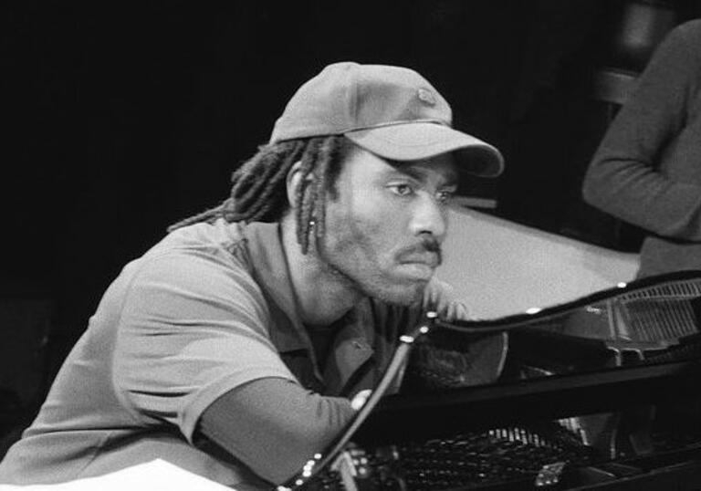 Black and white image of Devonte, leaning pensively on a piano wearing a baseball cap.