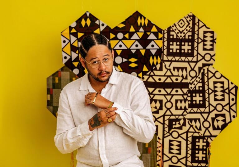 Image of Emicida posing in front of a bright yellow wall with a central black pattern