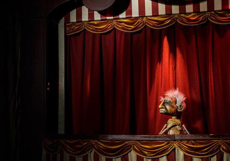 A rather sad looking puppet stands in a booth with a sign above their head that reads 'existential anguish'.