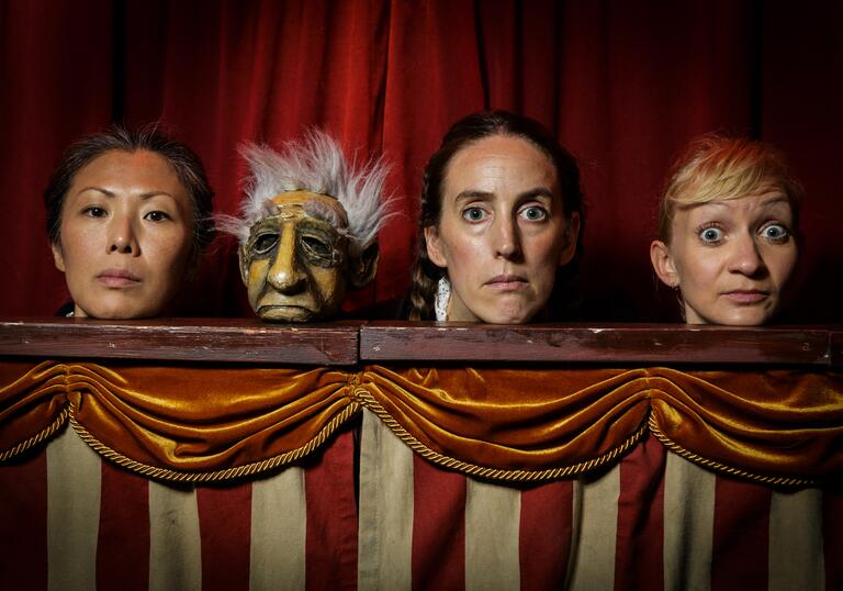 Three performers and one puppet stand in a puppet booth and stare directly at the camera.