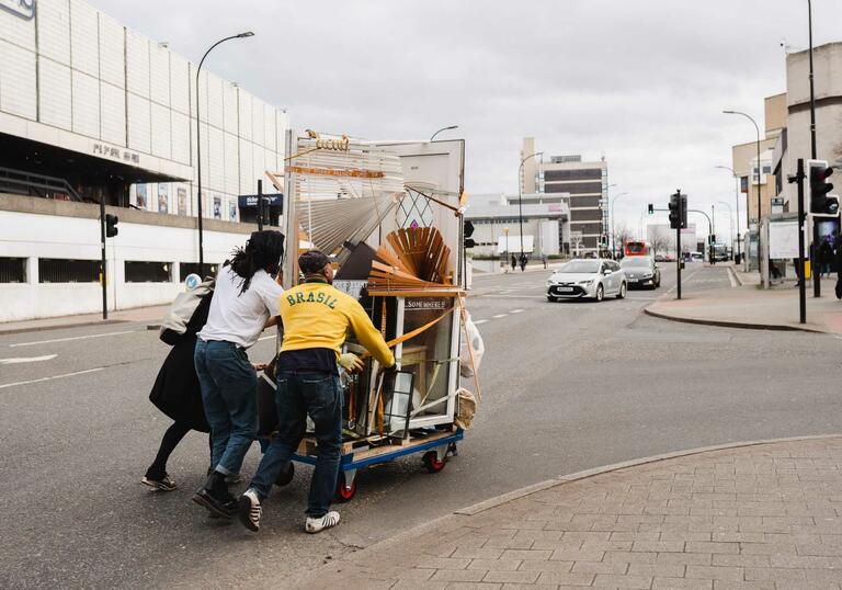 image of three people pushing a trolley with furniture up a hill with buildings and cars in the background