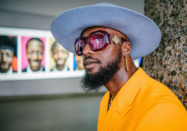 Kay is a black male, wearing a baby blue hat and yellow suit. He is standing in front of the S.M.I.L.E-ing Boys Experience installation