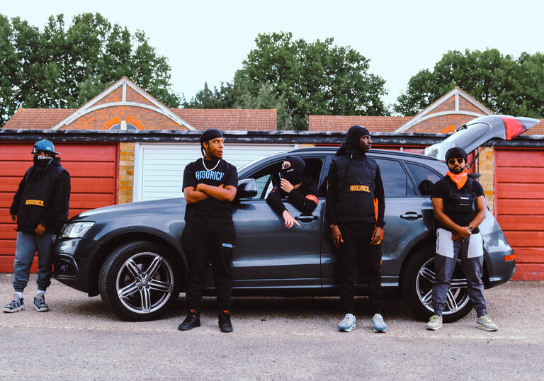 Performers of The UK Drill Project stand around a car. 