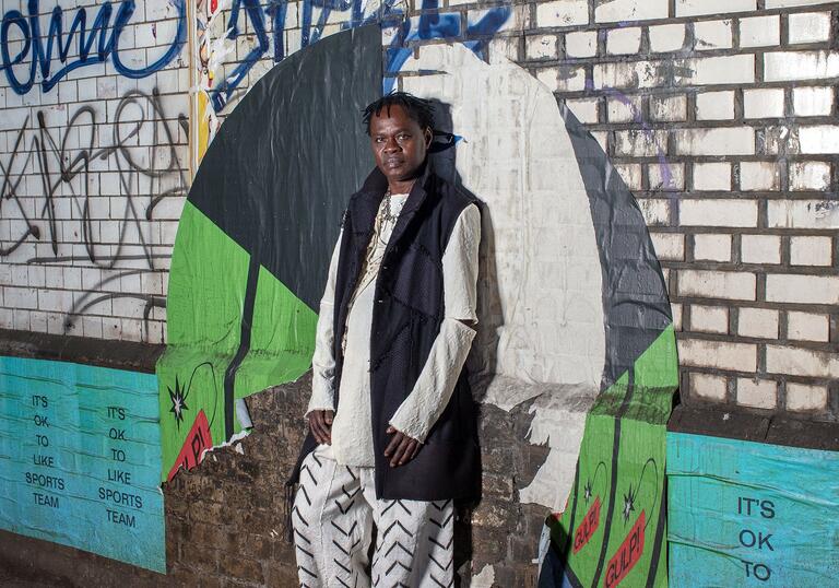 Dynamic image of Baaba Maal, standing against a graffiti covered brick wall