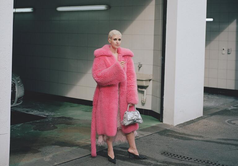 Dillon photographed in an urban parking lot wearing a pink furry coat in heels, and a cigarette in her hand