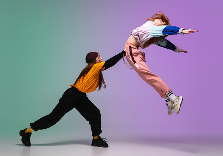 Two girls dancing, one with arm outstretched, pushing the other into the air, against a pastel coloured background