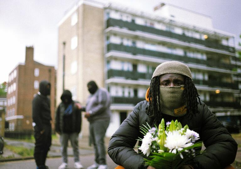 A performer wearing a face covering and holding a bunch of white flowers crouches down in front of a block of flats. Behind them a group of 3 other performers stand talking.