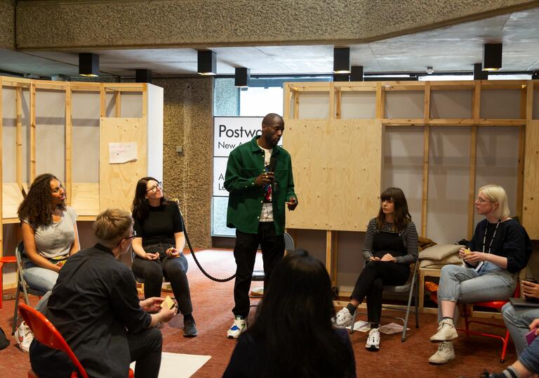 Six people sat on chairs in a circle watch as one man who is standing with a microphone leads the discussion