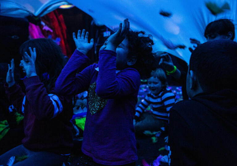 A group of children gather underneath a large blue sheet. They laugh, smile and lift their hairs upwards towards it.