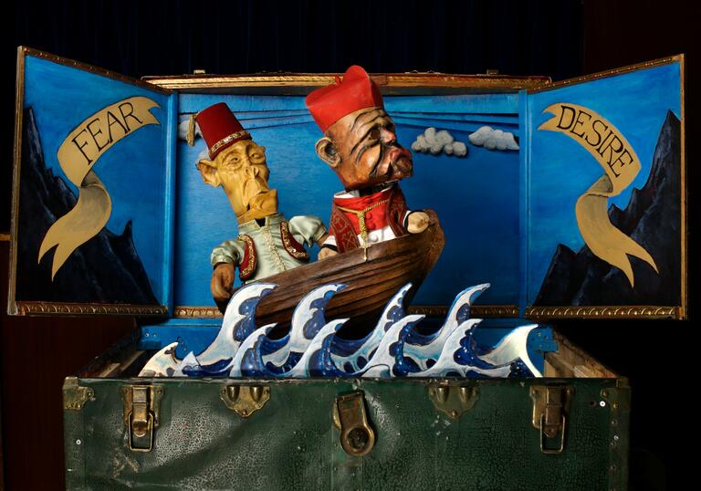 Two small puppets wearing red hats are sailing against waves on a boat inside a travel case. The words fear and desire are written on the lid.