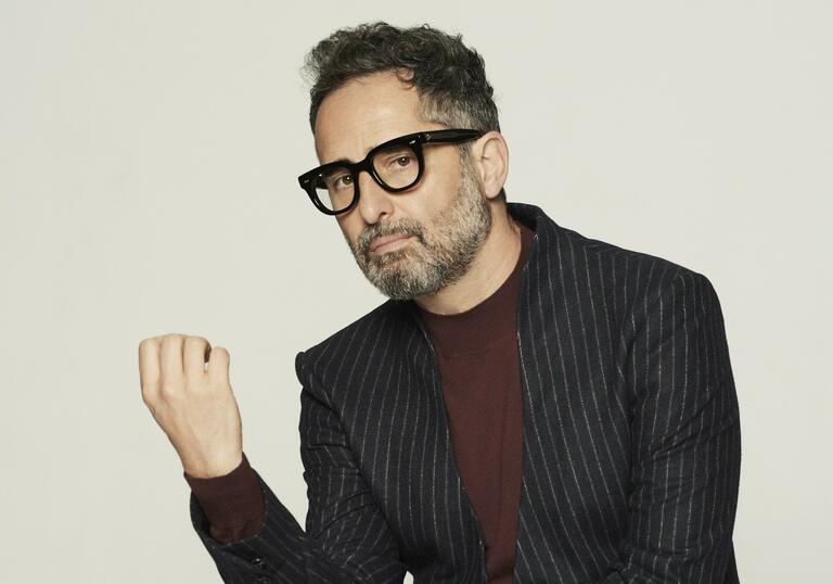 Press shot of Jorge Drexler in a pinstripe suit jacket, burgundy shirt and black rimmed glasses, starring into the camera