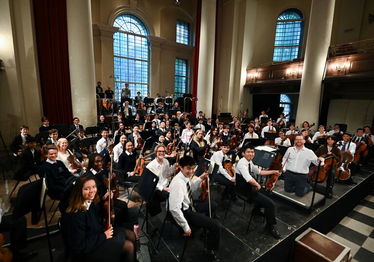 The Westminster Symphony Orchestra sitting on stage holding their instruments while turning to look at the camera