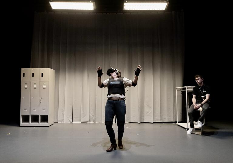 A person wearing a VR headset stands with their arms in the air as if backing away from something. They stand in front of a cream curtain and, to the right-hand side, somebody sits watching them as if supervising.