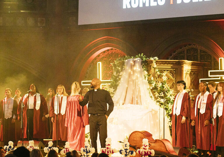 A choir performing on a stage with set pieces including a Christian cross in neon yellow and a bed draped in sheer canopy with trailing flowers