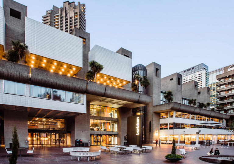 the barbican at evening with yellow lights on and a few people sitting on white benches outside