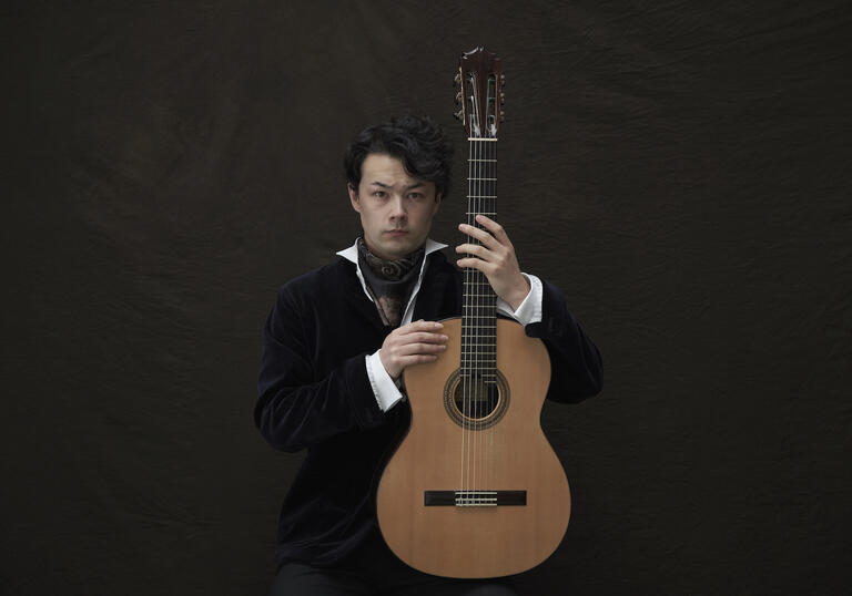 Sean Shibe sitting in front of a dark grey background, holding his guitar upright