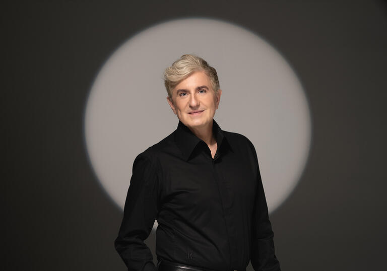 Jean Yves Thibaudet standing in front of a grey background with a spotlight forming a light circle behind him